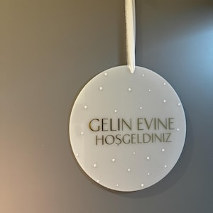 May include: A round, white sign with the text "GELIN EVINE. HOSGELDINIZ" in gold lettering. The sign is decorated with small, scattered, white dots and is suspended by a white ribbon. The sign is hanging on a gray wall.