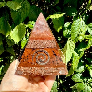 May include: A pyramid-shaped object made of clear resin containing layers of reddish-brown stone fragments and copper shavings. A metallic spiral symbol is embedded on the front face of the pyramid, which features a smooth, polished finish.