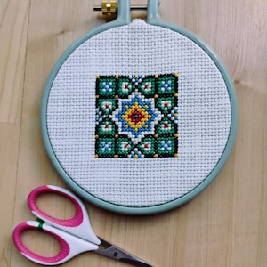 May include: A completed cross-stitch project in a round, light blue embroidery hoop. The cross-stitch design features a geometric pattern with blue, green, yellow, and red colors. A pair of white and pink handled scissors rests below the hoop.