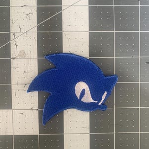 以下が含まれることがあります： 人気のゲームキャラクターの頭部が描かれた青色の刺繍パッチ。パッチには白い目と口のディテールがあります。パッチは、黒とグレーのチェック柄の表面に配置されています。