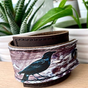 May include: A brown leather cuff bracelet with a white background and a blue bird design. The bird is perched on a branch with a wood grain pattern.