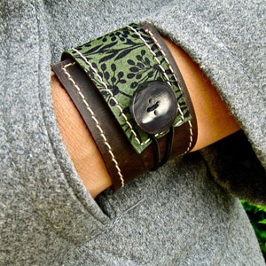 May include: A brown leather cuff bracelet with a green floral fabric overlay and a large black button. The bracelet is adjustable with a black cord.
