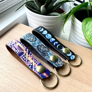 May include: Three leather keychains with different patterns. The first keychain has a blue and pink geometric pattern, the second has a blue and white mandala pattern, and the third has a black and blue moon phase pattern.