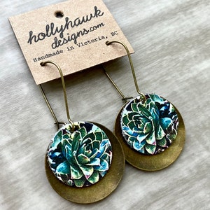 May include: A pair of brass earrings with a round, green and blue floral design. The earrings are handmade in Victoria, BC.