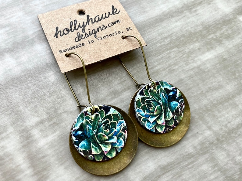 May include: A pair of brass earrings with a round, green and blue floral design. The earrings are handmade in Victoria, BC.
