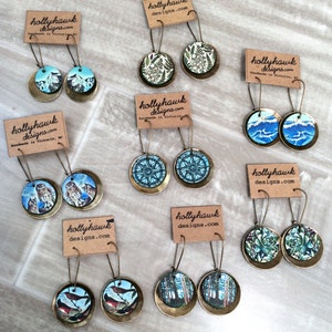 May include: Assortment of handmade earrings featuring various designs, including owls, birds, trees, and compass roses. The earrings are made with brass and have a vintage aesthetic. The earrings are displayed on a light brown surface with small cards that say "hollyhawk designs.com".