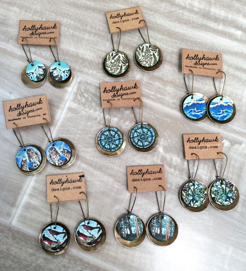 May include: Assortment of handmade earrings featuring various designs, including owls, birds, trees, and compass roses. The earrings are made with brass and have a vintage aesthetic. The earrings are displayed on a light brown surface with small cards that say "hollyhawk designs.com".