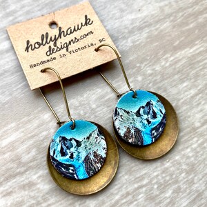May include: A pair of dangle earrings featuring a round, blue and white mountain scene on a brass disc. The earrings are handmade in Victoria, BC.