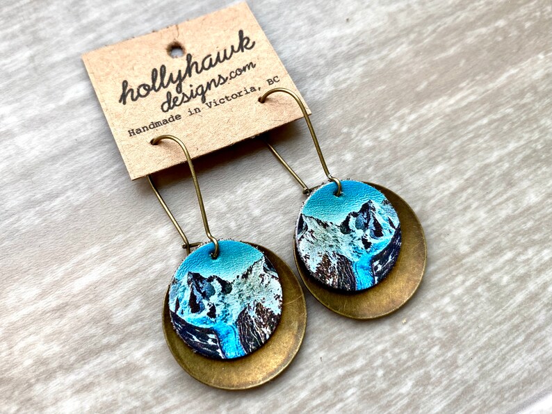 May include: A pair of dangle earrings featuring a round, blue and white mountain scene on a brass disc. The earrings are handmade in Victoria, BC.