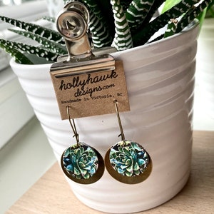 May include: A pair of gold tone earrings with a round, green and blue succulent design. The earrings are hanging from a white pot with a plant inside. The pot has a brown paper tag attached to it that reads "hollyhawk designs.com Handmade in Victoria, BC".