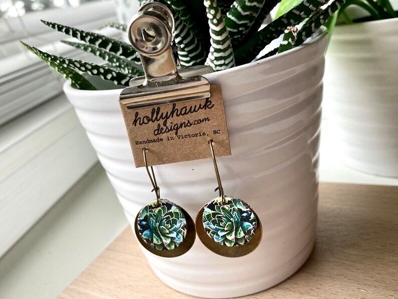May include: A pair of gold tone earrings with a round, green and blue succulent design. The earrings are hanging from a white pot with a plant inside. The pot has a brown paper tag attached to it that reads "hollyhawk designs.com Handmade in Victoria, BC".