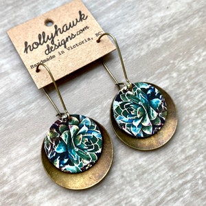 May include: A pair of brass and floral patterned earrings. The earrings are round and have a layered design. The floral pattern is green, blue, and white.