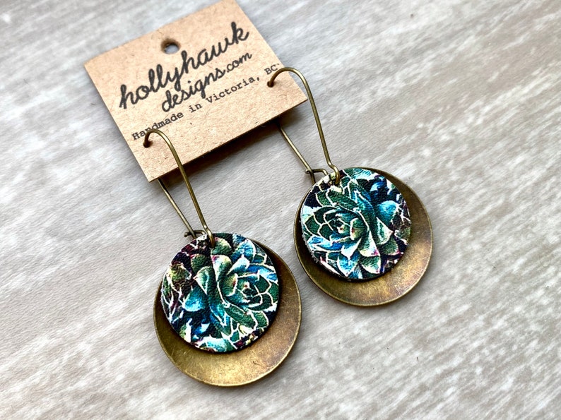 May include: A pair of brass and floral patterned earrings. The earrings are round and have a layered design. The floral pattern is green, blue, and white.
