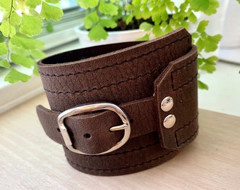 May include: A brown leather cuff bracelet with a silver buckle and two silver rivets. The bracelet has a stitched design.
