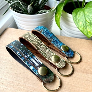 May include: Three leather keyrings with different designs. One keyring features a blue and white compass design, another has a green and white floral pattern, and the third has a forest scene. Each keyring has a gold-coloured metal ring.