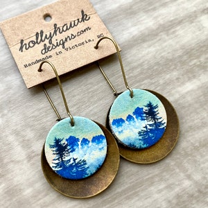 May include: A pair of dangle earrings with a blue and white mountain scene on a round, brass-colored base. The earrings are handmade in Victoria, BC by hollyhawkdesigns.com.
