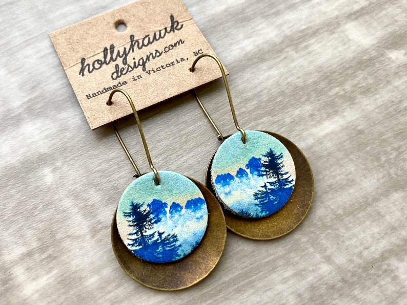 May include: A pair of dangle earrings with a blue and white mountain scene on a round, brass-colored base. The earrings are handmade in Victoria, BC by hollyhawkdesigns.com.