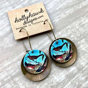 May include: A pair of dangle earrings with a round, brass-colored metal base and a blue and brown patterned fabric top featuring a vintage illustration of birds. The earrings are handmade in Victoria, BC by hollyhawkdesigns.com.
