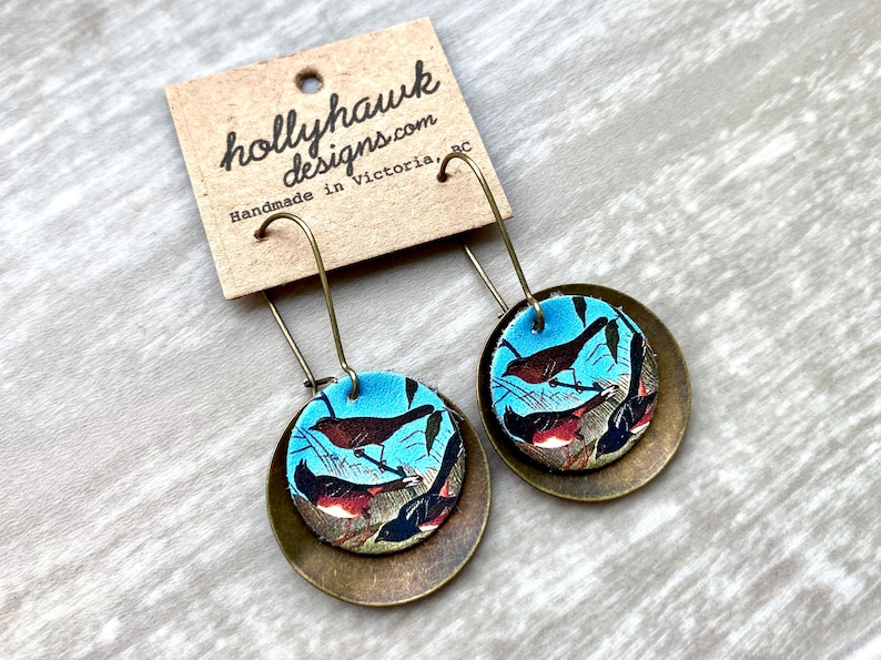 May include: A pair of dangle earrings with a round, brass-colored metal base and a blue and brown patterned fabric top featuring a vintage illustration of birds. The earrings are handmade in Victoria, BC by hollyhawkdesigns.com.