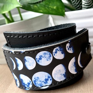 May include: A black leather bracelet with a moon phase pattern. The bracelet is adjustable and has a silver snap closure.