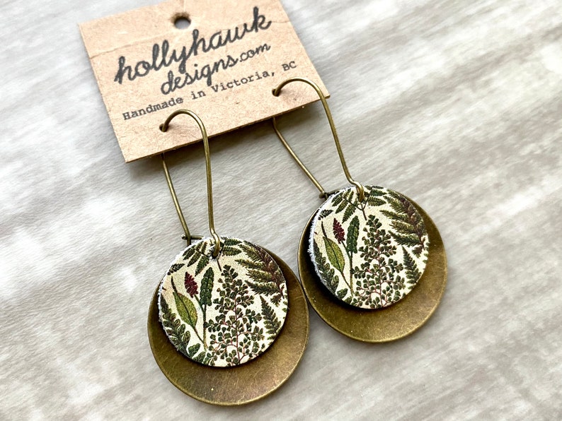 May include: A pair of gold tone earrings with a round, white and green floral pattern on the front. The earrings are handmade in Victoria, BC by hollyhawkdesigns.com.