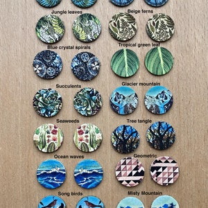 May include: A set of 12 pairs of round, colorful, cork-backed earrings with various nature-inspired designs. The designs include turquoise ferns, compass, jungle leaves, beige ferns, tropical green leaf, blue crystal spirals, glacier mountain, succulents, seaweeds, tree tangle, ocean waves, geometric, song birds, and misty mountain.