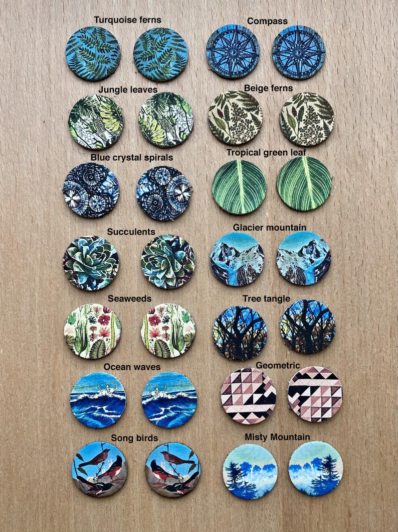 May include: A set of 12 pairs of round, colorful, cork-backed earrings with various nature-inspired designs. The designs include turquoise ferns, compass, jungle leaves, beige ferns, tropical green leaf, blue crystal spirals, glacier mountain, succulents, seaweeds, tree tangle, ocean waves, geometric, song birds, and misty mountain.