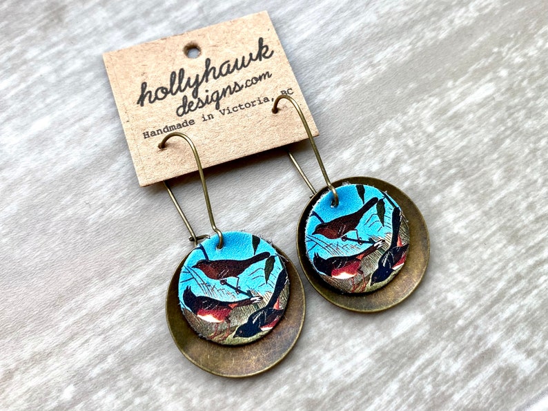 May include: A pair of brass earrings with a blue and brown bird print design. The earrings are handmade in Victoria, BC by hollyhawkdesigns.com.