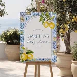 May include: A wooden easel displays a baby shower sign. The sign features a white background with blue and yellow tile-like borders and lemon illustrations. Text reads "Welcome to Isabella's baby shower" and the date.