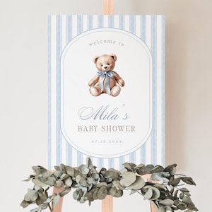 May include: A baby shower welcome sign with a light blue striped background. The sign features a watercolour illustration of a teddy bear with a blue bow. Text reads "Welcome to Mila's Baby Shower" and the date.