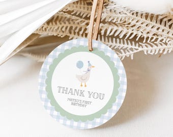 May include: Round blue and white gingham thank you tag with a green circle in the center. The green circle features a cartoon duck wearing a party hat and holding a blue balloon. The text "Thank You" is in a white font with a black outline. Below the text is "Mateo's First Birthday" in a smaller font.
