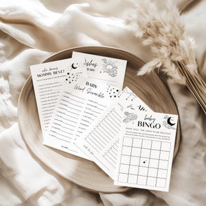 Könnte beinhalten: Eine Auswahl an Babyparty-Spielkarten mit schwarzem Text und Illustrationen auf weißem Hintergrund. Die Karten sind auf einem Holztablett angeordnet. Spiele umfassen "Wer kennt Mama am besten?", "Baby-Wortsalat" und "Baby-Bingo".