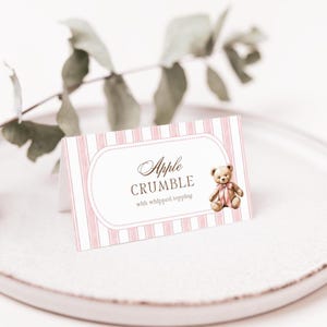 May include: A pink and white striped table card with the text "Apple Crumble with whipped topping" in elegant script. A small teddy bear illustration with a pink bow is on the right. The card is on a white plate.