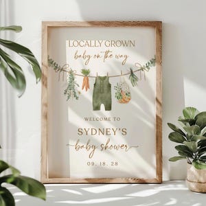 May include: A white sign with a brown wooden frame announcing a baby shower. The sign says "Locally Grown baby on the way" with a green baby onesie hanging on a clothesline with vegetables and herbs. The sign also says "Welcome to Sydney's baby shower 09.18.28".