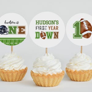 Puede incluir: Tres adornos para cupcakes con tem&aacute;tica de f&uacute;tbol americano. Los adornos presentan un casco de f&uacute;tbol, un bal&oacute;n de f&uacute;tbol y el texto "hudson is one", "hudson's first year down" y "1".