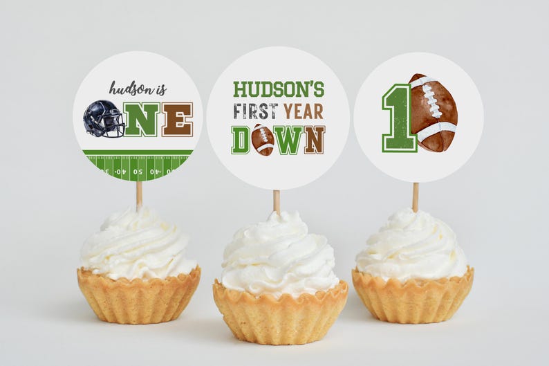 Puede incluir: Tres adornos para cupcakes con tem&aacute;tica de f&uacute;tbol americano. Los adornos presentan un casco de f&uacute;tbol, un bal&oacute;n de f&uacute;tbol y el texto "hudson is one", "hudson's first year down" y "1".