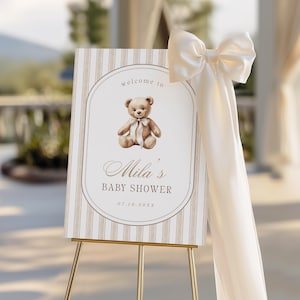 Pode incluir: Uma placa de boas-vindas para chá de bebê com uma ilustração de urso de pelúcia. A placa tem um fundo listrado bege e branco com o texto "welcome to Mila's BABY SHOWER" e a data 07.10.20XX. Um grande laço creme adorna a lateral.