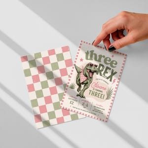 May include: A rectangular birthday invitation card featuring a green T-Rex wearing pink heart-shaped sunglasses and a checkered bandana. The text reads three REX, Noane is turning THREE, and party details. A matching pink and green checkered card is included.