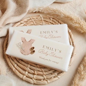 Op de afbeelding: Twee witte chocolade reep verpakkingen met een illustratie van een roze baby romper en bruine schoentjes. De tekst op de verpakking luidt "Emily's Baby Shower" en "Bedankt voor je komst!"