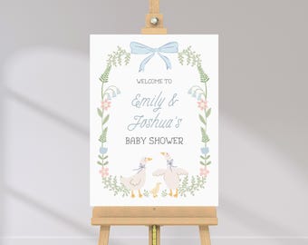 Könnte beinhalten: Ein Babyparty-Schild auf einer Holzstaffelei. Auf dem Schild steht "Willkommen zu Emilys & Joshuas Babyparty" in eleganter Schrift. Das Design umfasst eine blaue Schleife, einen Blumenkranz und Illustrationen von Gänsen und einem Entlein.
