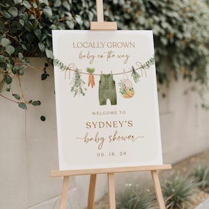 May include: A white sign with brown text announcing a baby shower for Sydney. The sign says "Locally Grown baby on the way" and features a green baby onesie hanging on a clothesline with vegetables and greenery. The date of the shower is 09.18.28.