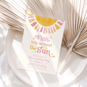 May include: A white invitation card with a yellow and pink sun design and the text "First trip around the Sun." The invitation is for Ella's First Birthday. The card is on a white plate with a dried palm leaf.