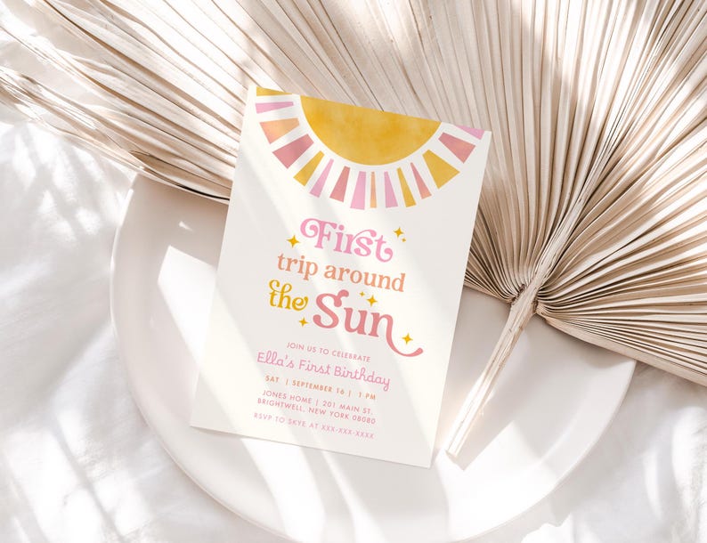 May include: A white invitation card with a yellow and pink sun design and the text "First trip around the Sun." The invitation is for Ella's First Birthday. The card is on a white plate with a dried palm leaf.