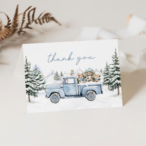 May include: A folded card featuring a watercolour illustration of a light blue vintage truck carrying woodland animals in a snowy scene. The words "thank you" are written in blue script across the top.