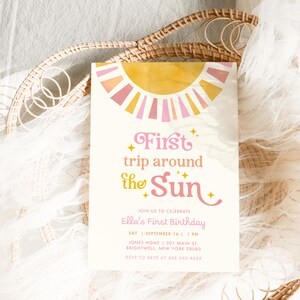 May include: A cream-colored invitation card with a sun design in pink, yellow, and orange. The text reads "First trip around the Sun" and announces Ella's First Birthday. The card includes date, time, and location details.