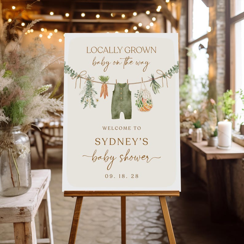 May include: A white sign with brown text announcing a baby shower for Sydney. The sign features a watercolor illustration of a baby onesie hanging on a clothesline with vegetables and greenery. The text reads "Locally Grown baby on the way" and "Welcome to Sydney's baby shower 09.18.28".