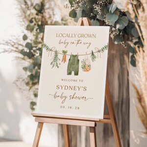 May include: A welcome sign for a baby shower with the text "Locally Grown baby on the way" and a green baby onesie hanging on a clothesline with vegetables and greenery. The sign also says "Welcome to Sydney's baby shower 09.18.28".
