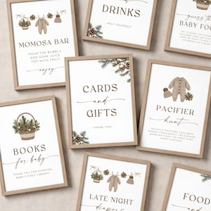 May include: A collection of framed signs for a baby shower, featuring brown text and watercolor illustrations of pine branches, pinecones, and baby clothing. The signs include titles like Momosa Bar, Cards and Gifts, Books for Baby, and Pacifier Hunt.