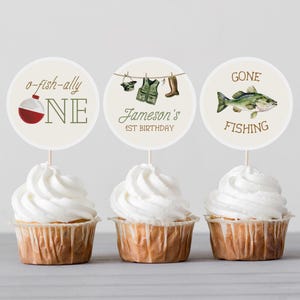 May include: Three white cupcakes with round, white toppers. The toppers have a fishing theme and say "o-fish-ally one", "Jameson's 1st Birthday", and "Gone Fishing".