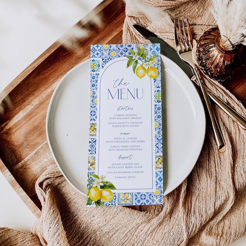 Puede incluir: Una tarjeta de men&uacute; con un dise&ntilde;o de azulejos azules y blancos, con limones y el texto "the MENU". El men&uacute; enumera entrantes, platos principales y postres. La tarjeta est&aacute; en un plato blanco.
